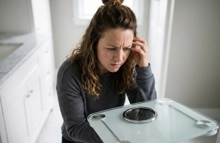 A frustrated woman looking down at a bathroom scale, illustrating unexplained weight gain as a hidden symptom of PCOS.
